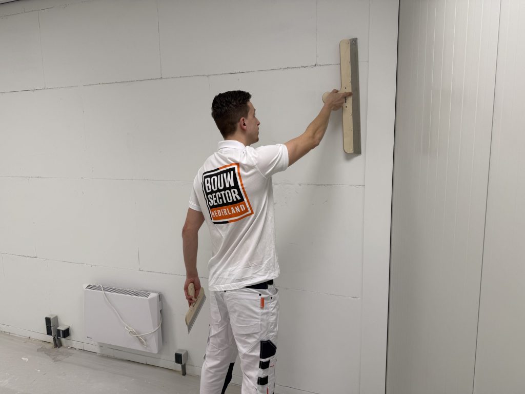 Stukadoor in wit werkpak werkt een lange wand af, met de spaan in zijn rechterhand en een plamuurspaan in zijn linker