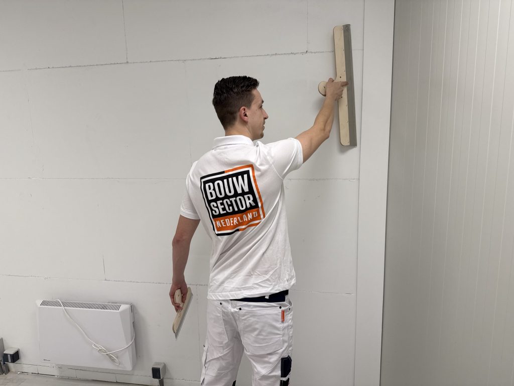 Stukadoor in een wit Bouwsector-shirt zet een lange spaan hoog tegen een wand van gipsblokken naast een elektrische kachel