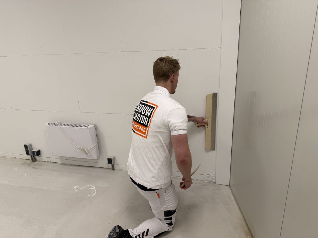 Knielende stukadoor strijkt met de spaan de pleister onderaan een wand glad, vlak bij de hoek van de kamer