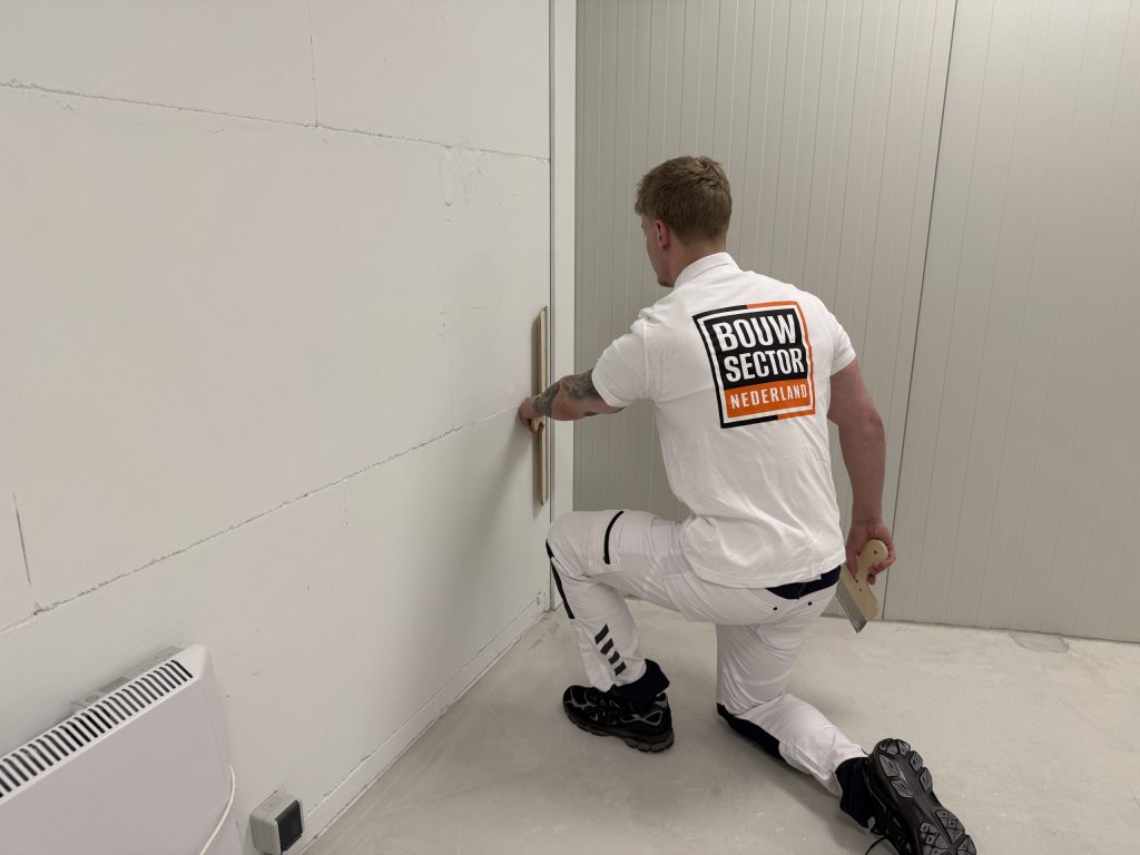 Stukadoor zit geknield bij een wand van gipsblokken en werkt met de spaan de onderkant bij de vloer af