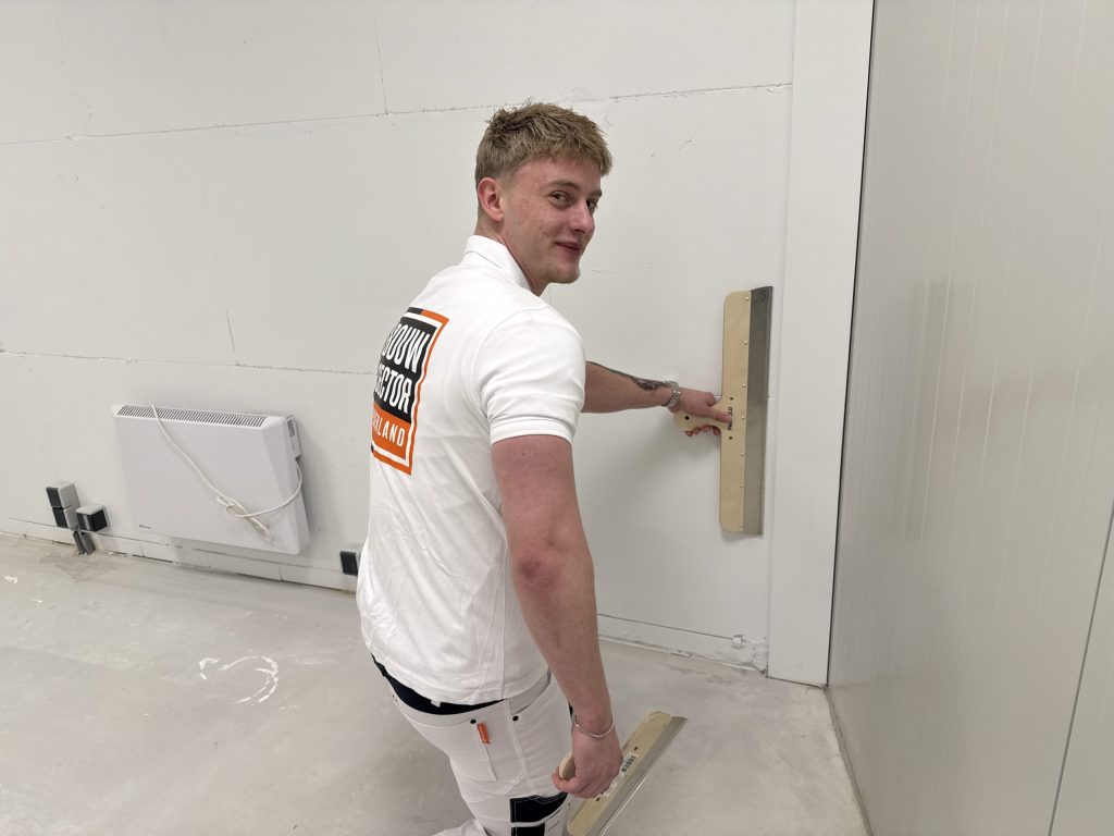 Stukadoor kijkt lachend om terwijl hij met een brede spaan de hoek van een wand afwerkt
