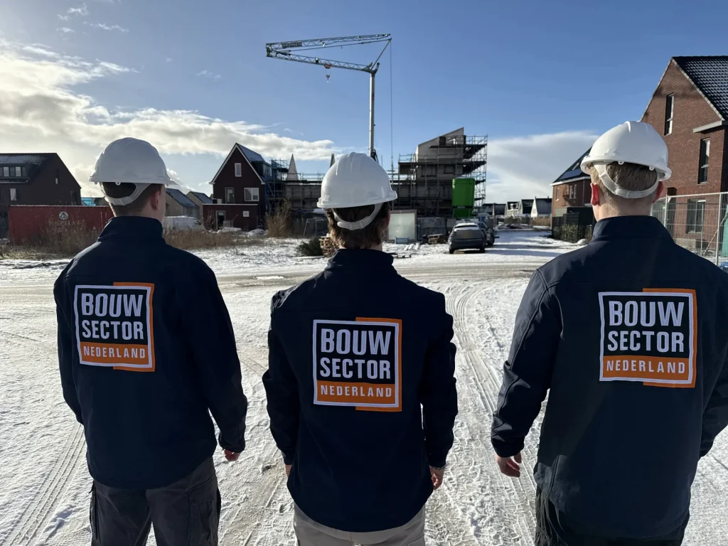 Drie vakmensen van Bouwsector Nederland met witte helm en donkerblauwe jas kijken uit over een nieuwbouwwijk met een bouwkraan