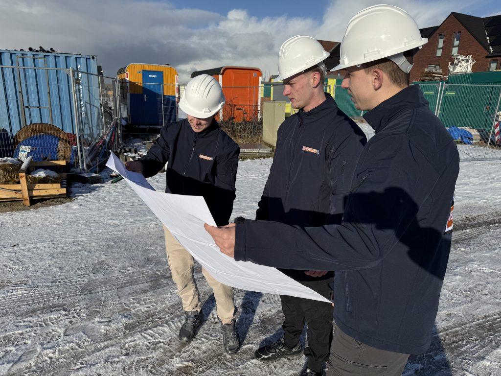 Drie vakmensen met witte helm bekijken samen een uitgeklapte tekening op een besneeuwde bouwplaats